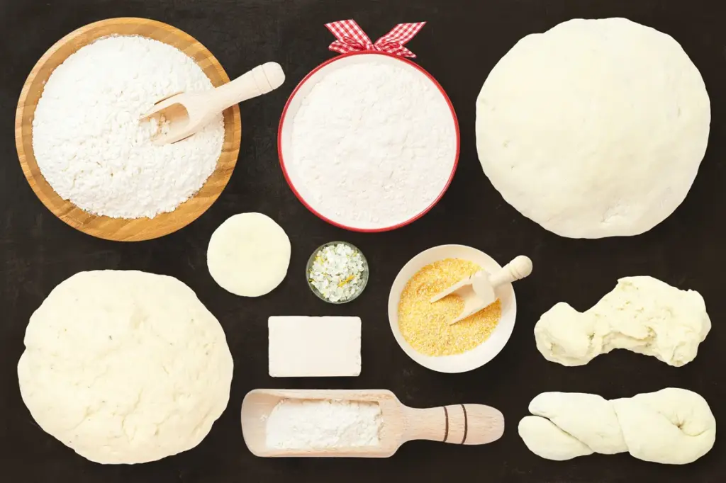 Bowls of flour, cornmeal, herbs, and various dough shapes arranged on a dark surface, showcasing baking ingredients and stages