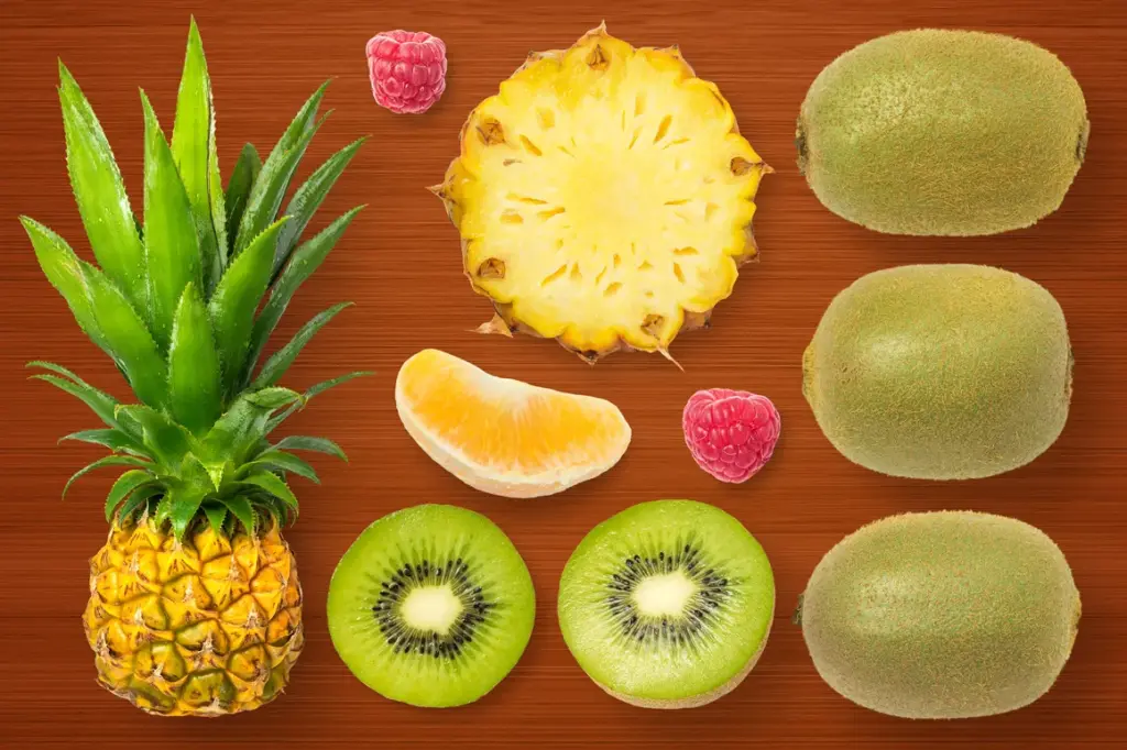 Whole and sliced pineapple, whole and halved kiwis, orange segment, and raspberries arranged on a wooden surface