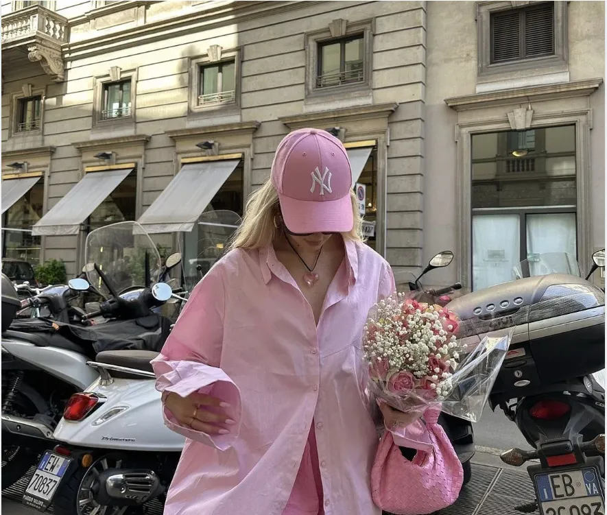 Stylish person in a pink outfit and New York Yankees hat holding a bouquet of flowers on a city street.